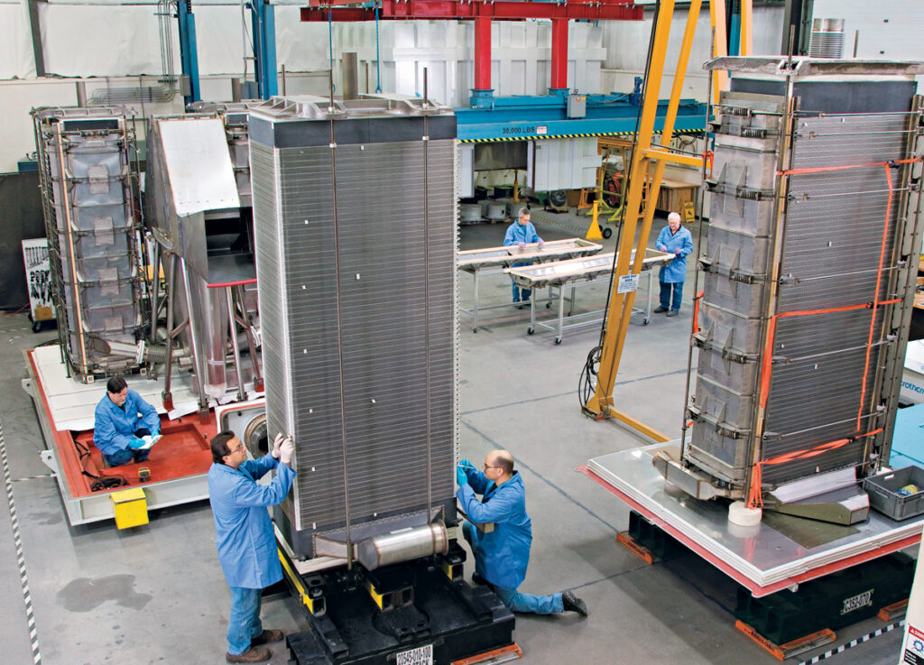 A group of people in blue coats work on tall mechanical things in a factory.